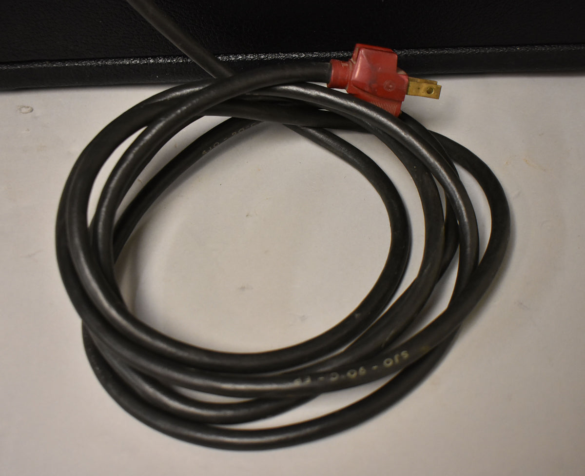 A coiled black power cable with a red, angled two-prong plug, resting on a white surface with a black amplifier edge visible.