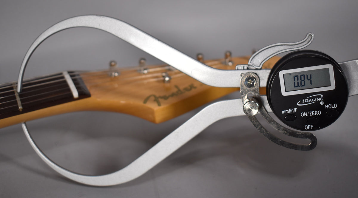 A digital caliper measures the neck thickness at the first fret, showing 0.84 inches, with the headstock and tuners blurred behind.