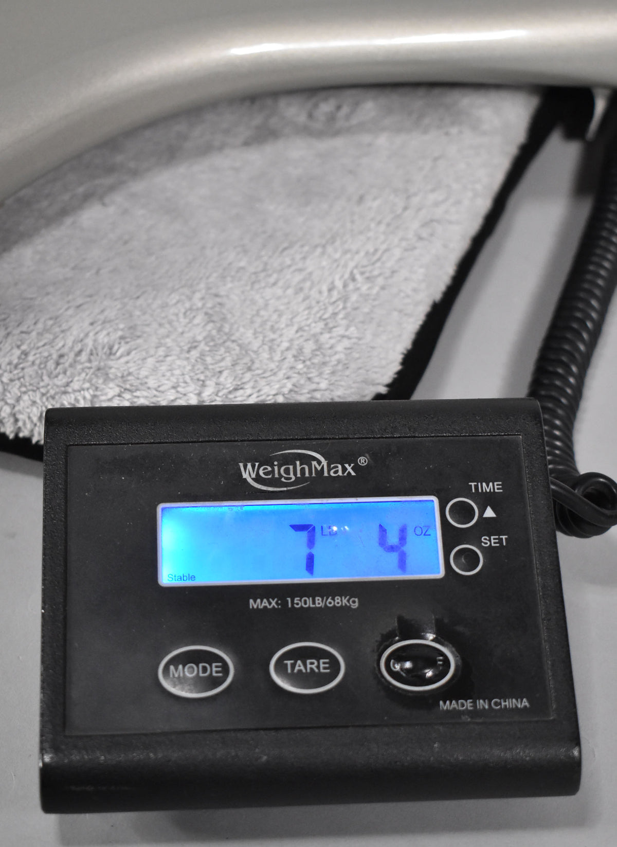 A close-up of a digital scale displaying "7 LB 4 OZ" in blue, with a soft grey blanket and a coiled black cable in the background.