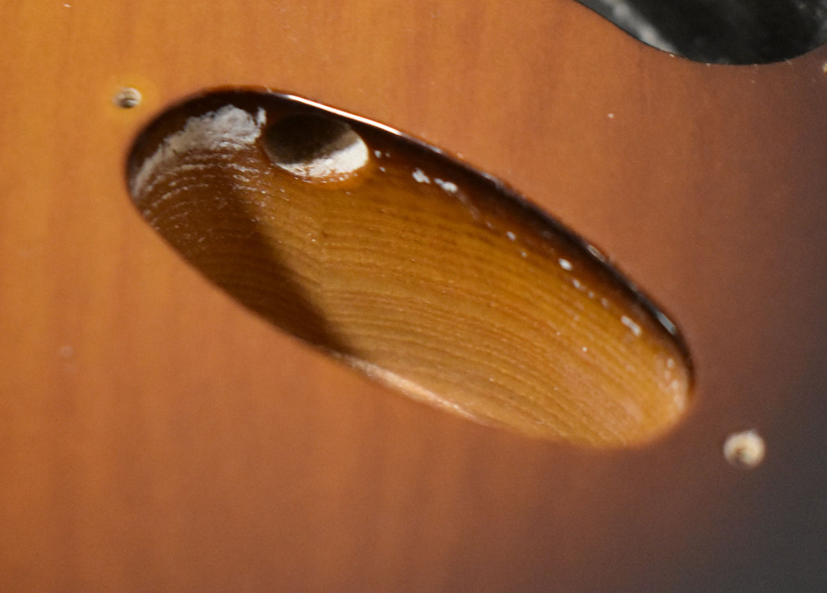 Close-up of the Stratocaster body's output jack cavity, showing wood grain and a glossy 2-tone sunburst finish with minor white specks.