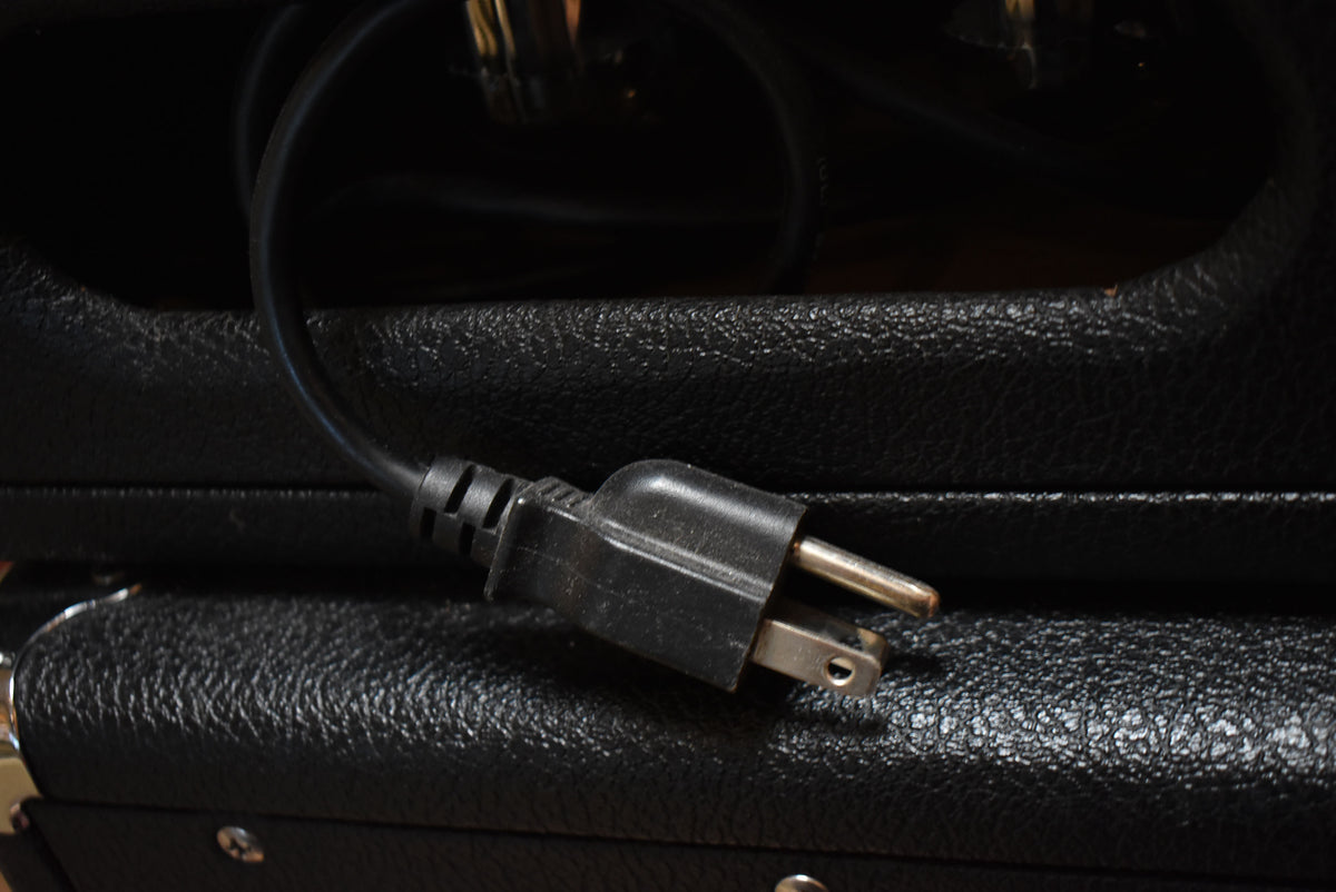 Close-up of a black power cord and 3-prong plug resting on the textured black tolex of the amp head, with some scuffs visible.