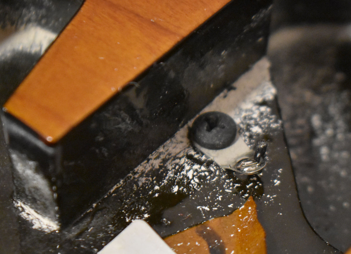 Close-up of the body's interior, showing a black screw and ground wire on a metal plate, surrounded by white dust and wood.