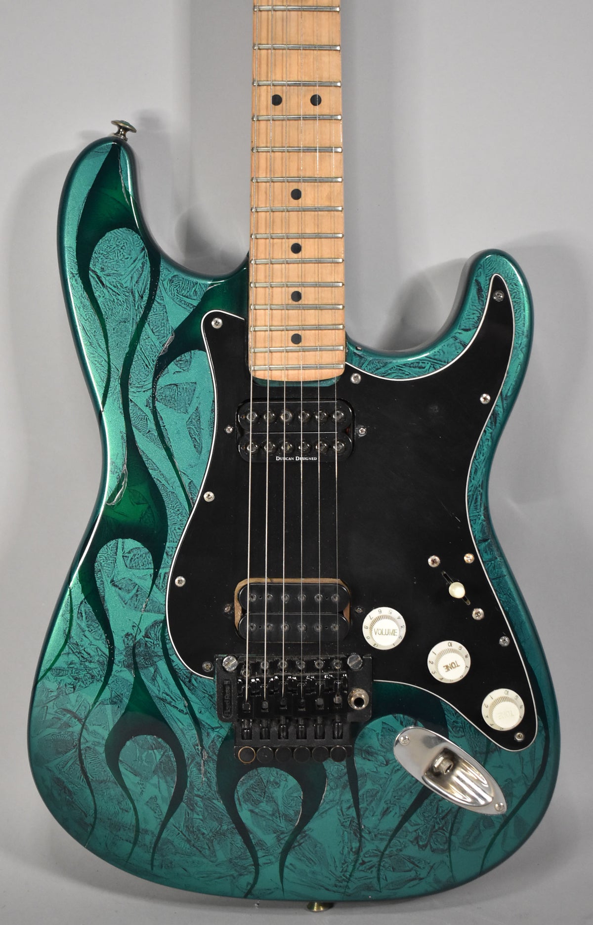 A green flame-finish electric guitar, angled slightly right, shows a maple fretboard, black pickguard, two humbuckers, and a Floyd Rose bridge.