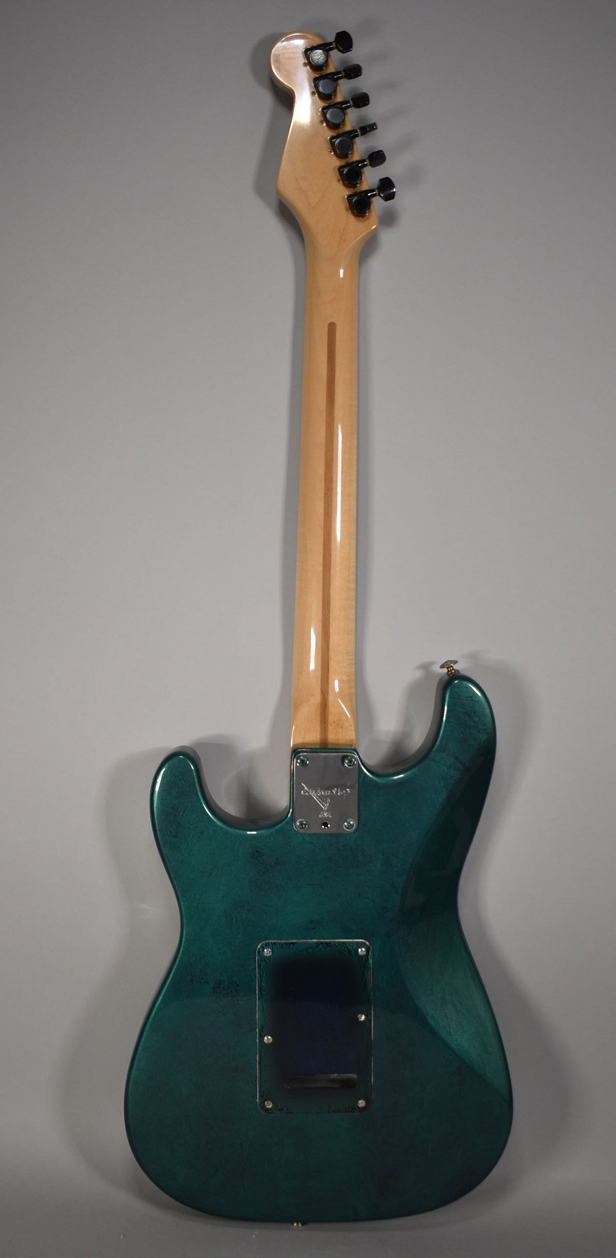 The back of a green flame finish Stratocaster, showing the maple neck, black tuners, and a rectangular tremolo cavity cover.