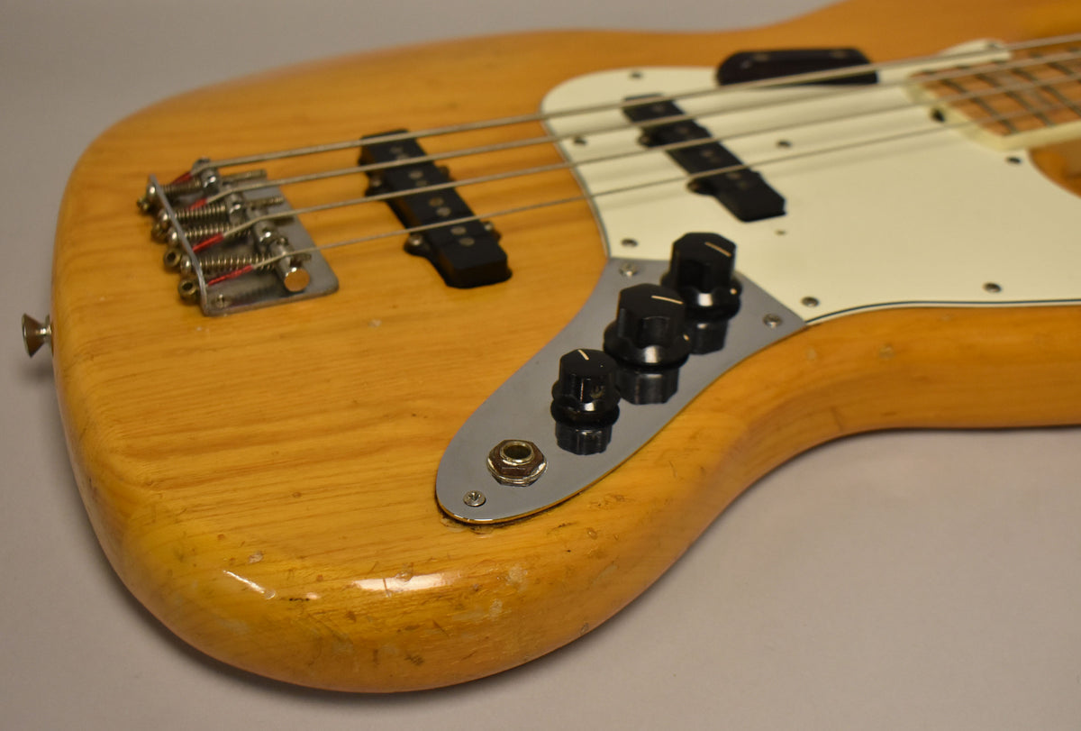 Close-up of the natural finish bass body, showing the bridge, pickups, control plate with three black knobs, and wear on the lower bout.
