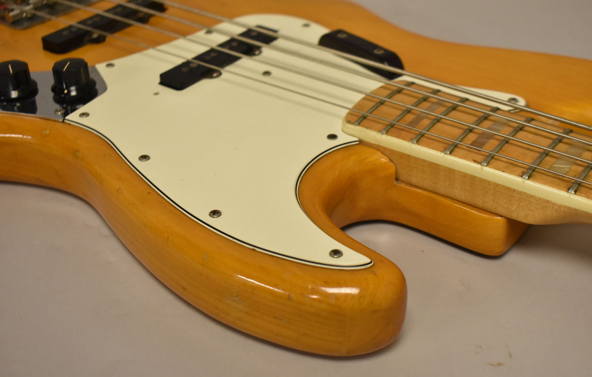 Close-up of a natural finish bass body with a white pickguard, two black pickups, and three black control knobs, showing minor wear.