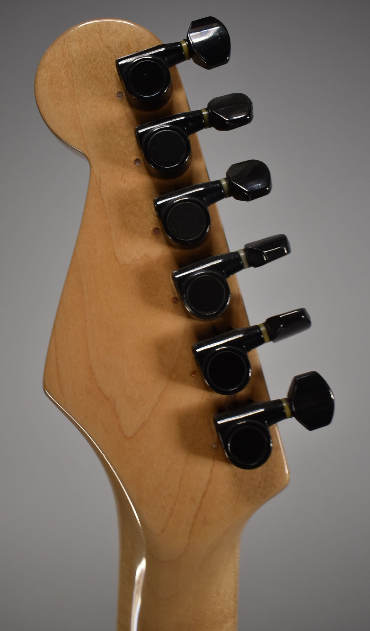 The back of a light wood headstock with six black tuning machines, angled slightly to the right.