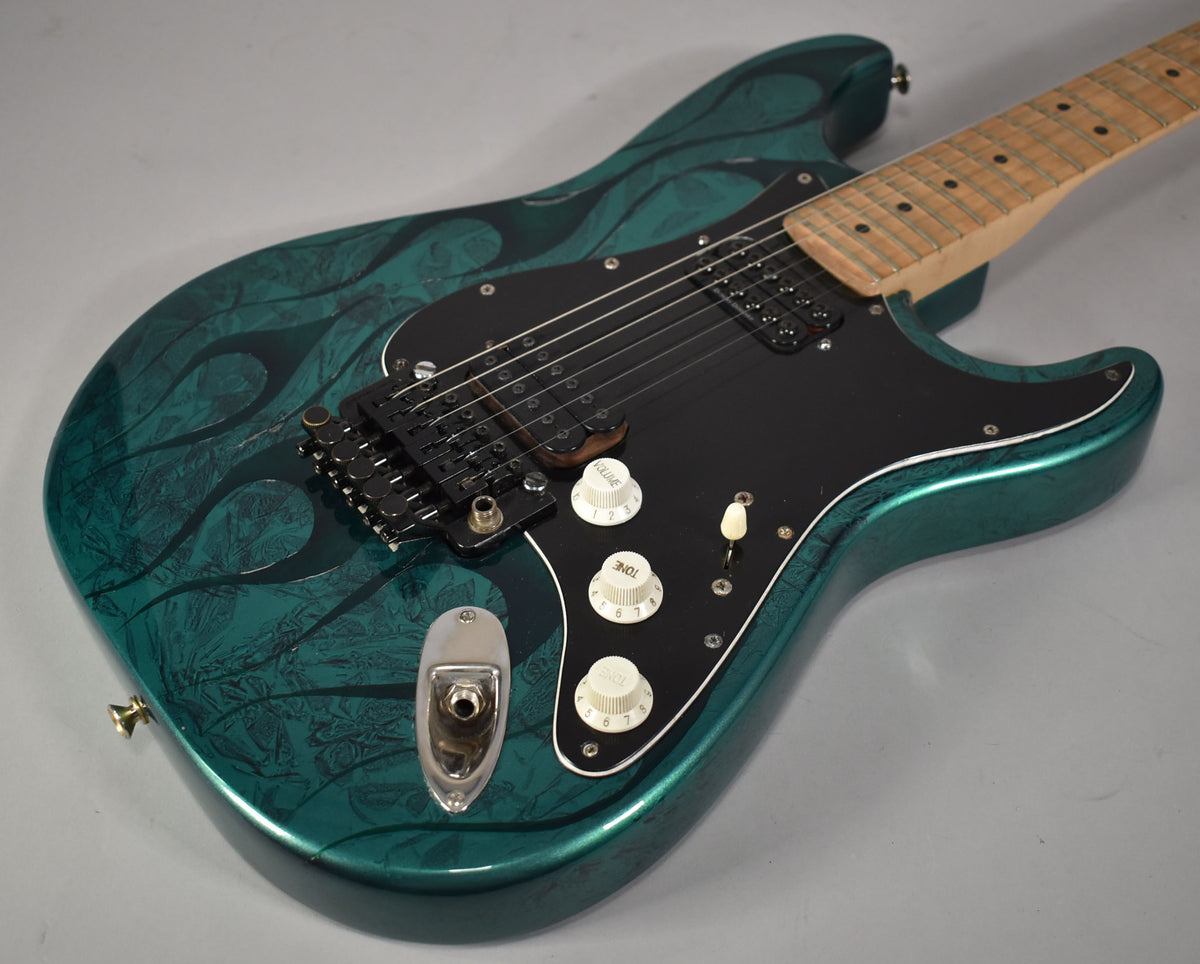 Angled view of a green flame-finish Stratocaster with a black pickguard, two humbuckers, three white knobs, and a black tremolo bridge.