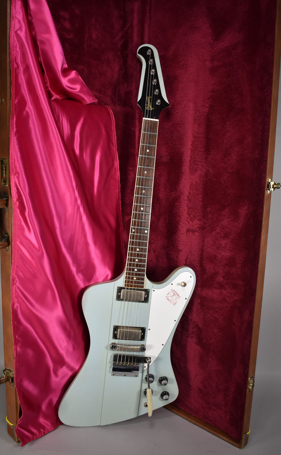 Full-length view of a pale blue electric guitar with a white pickguard featuring a red bird logo, resting in a red velvet-lined case.