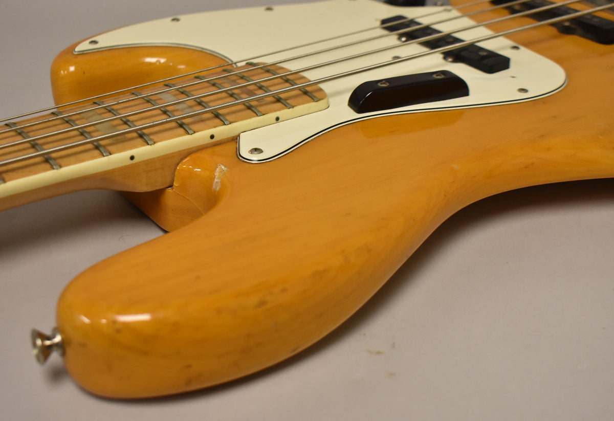 Upper body of a natural finish bass, showing a white pickguard, black pickups, and wear on the lower horn.