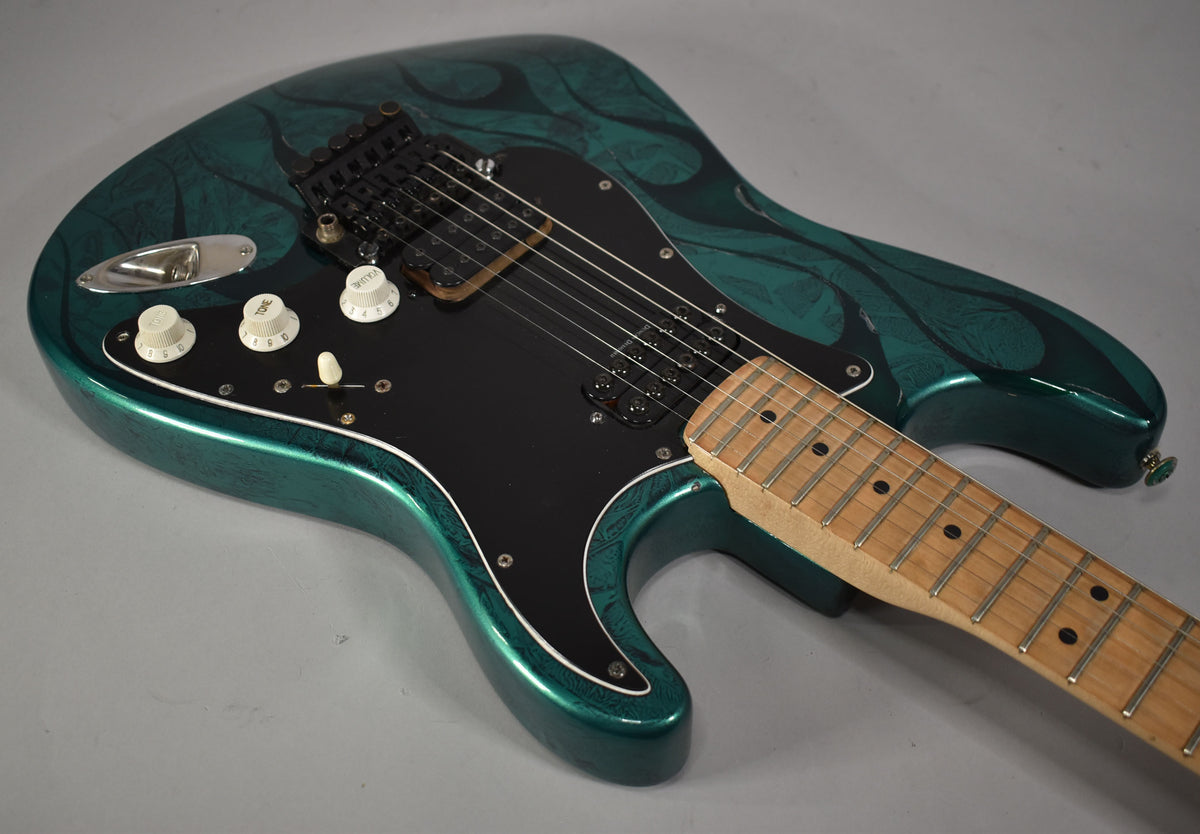 The green flame Stratocaster's body and neck are angled, showing the black pickguard, two humbuckers, and three white control knobs.