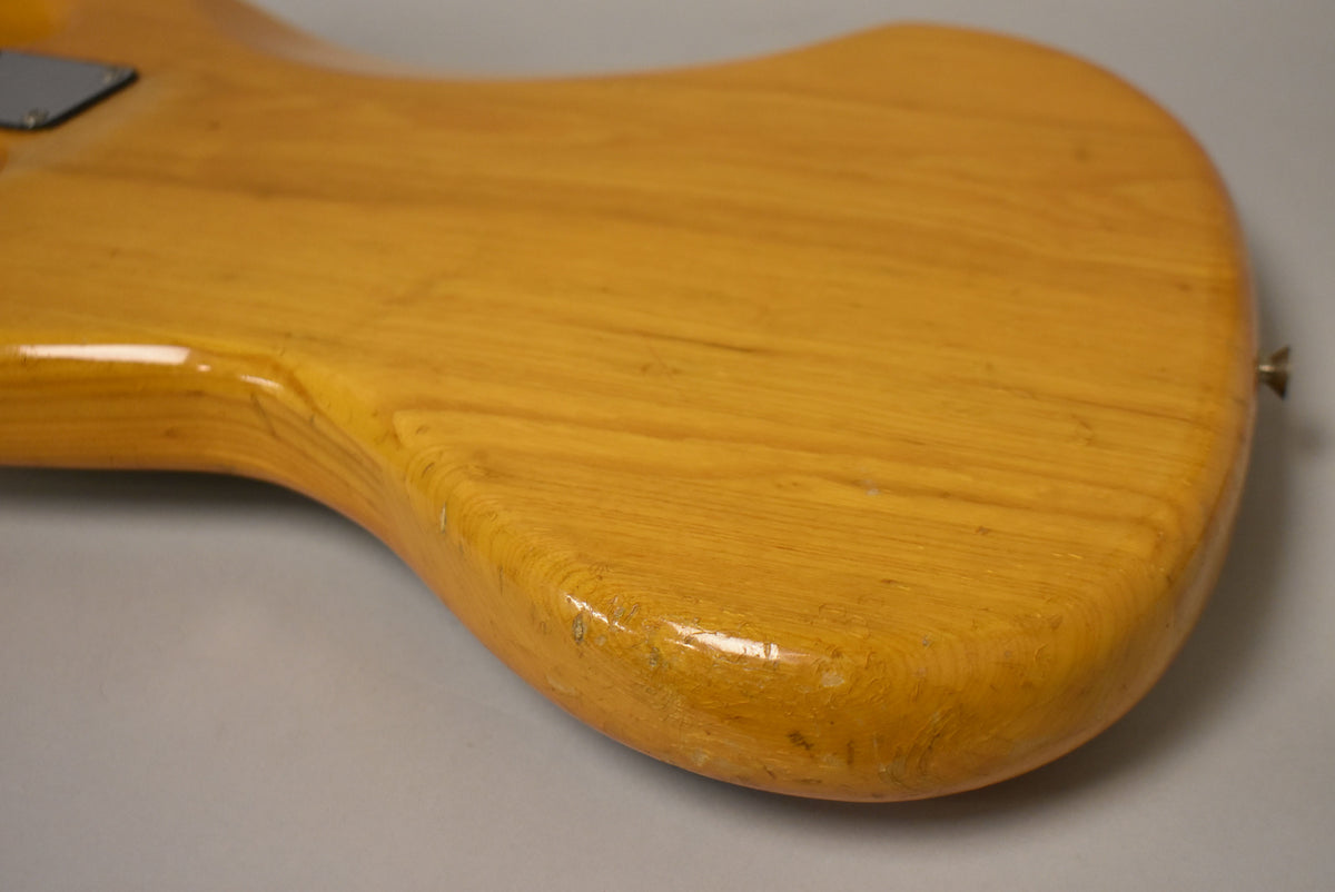 The lower bout of a natural finish bass guitar, showing finish wear, checking, and dings along the curved edges.