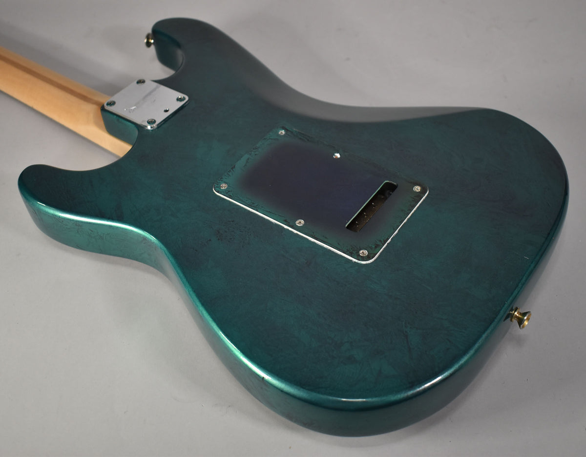 The back of a green flame finish Stratocaster body, showing a black tremolo cavity cover and a gold strap button.