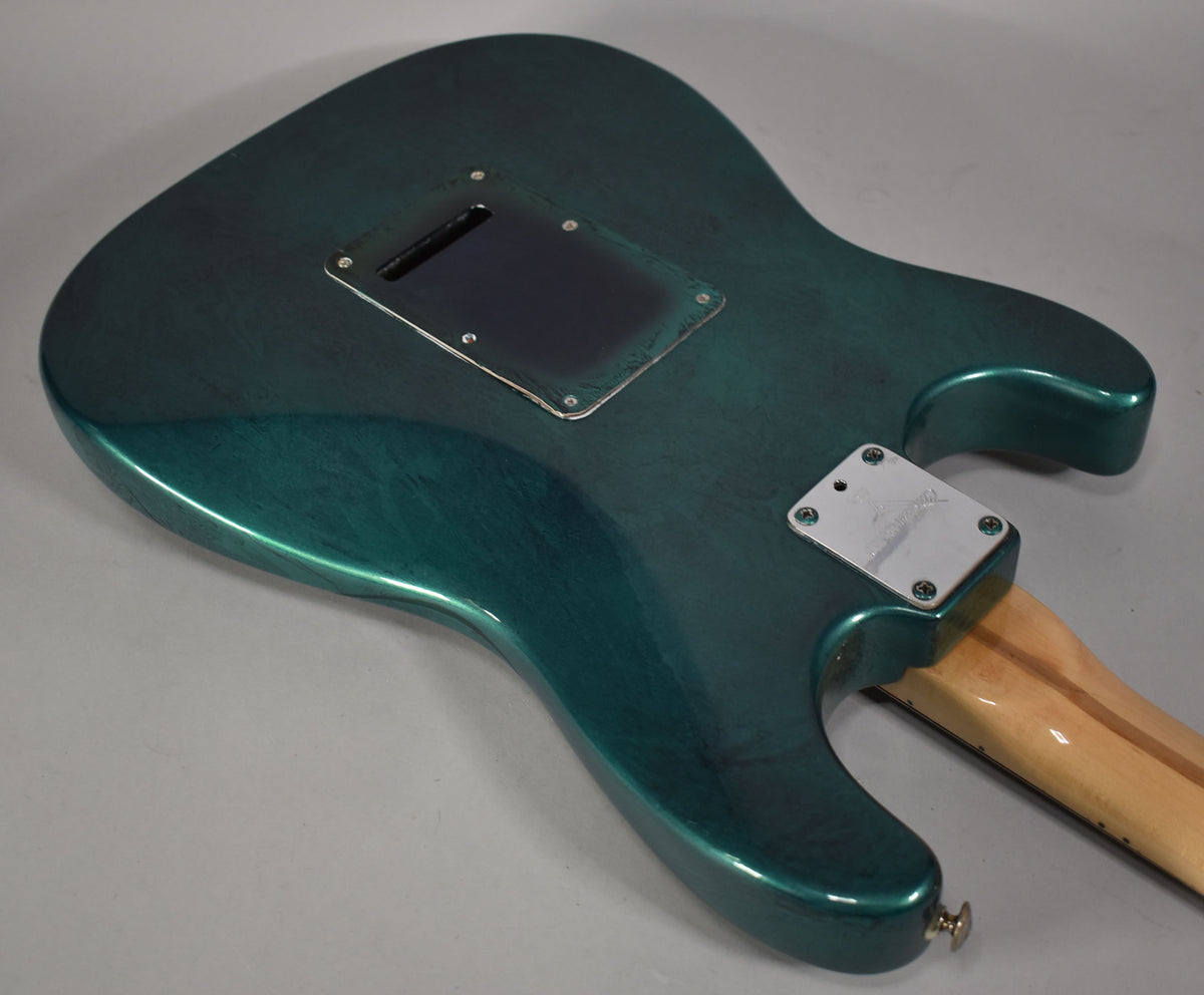 The back of a green flame finish Stratocaster body, showing the tremolo cavity cover, neck plate, and strap button.