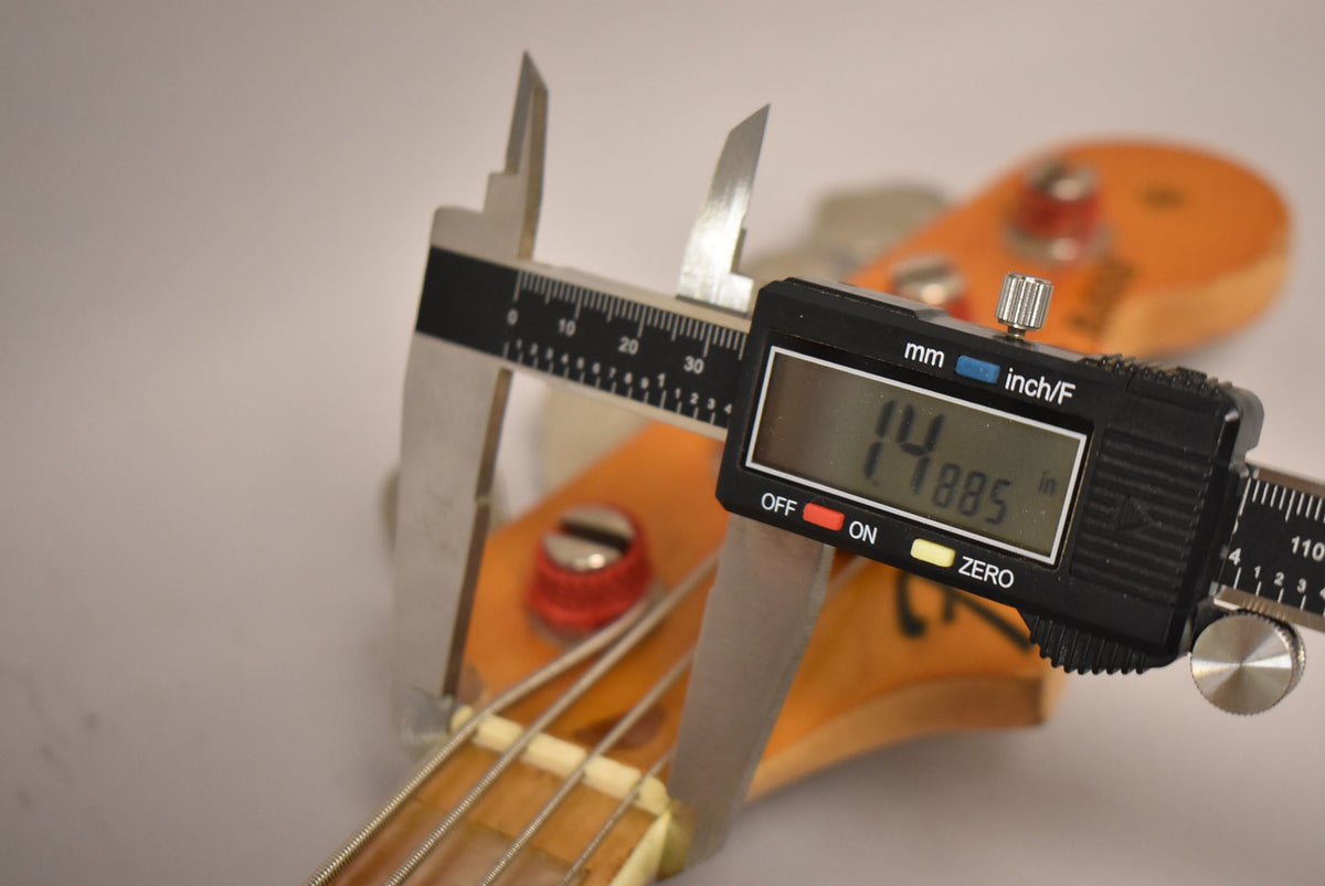 A digital caliper measures the bass's nut width at 1.4885 inches, with the headstock and tuning pegs visible.