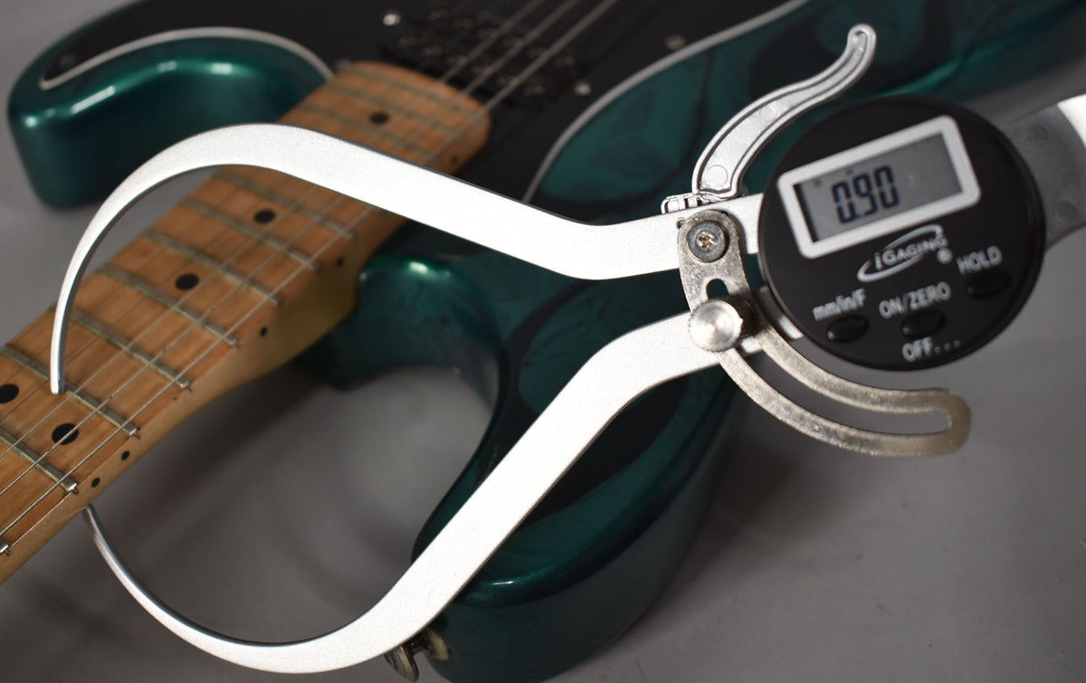 A digital caliper measures the fret height on the maple neck of the green flame Stratocaster, displaying "0.90".
