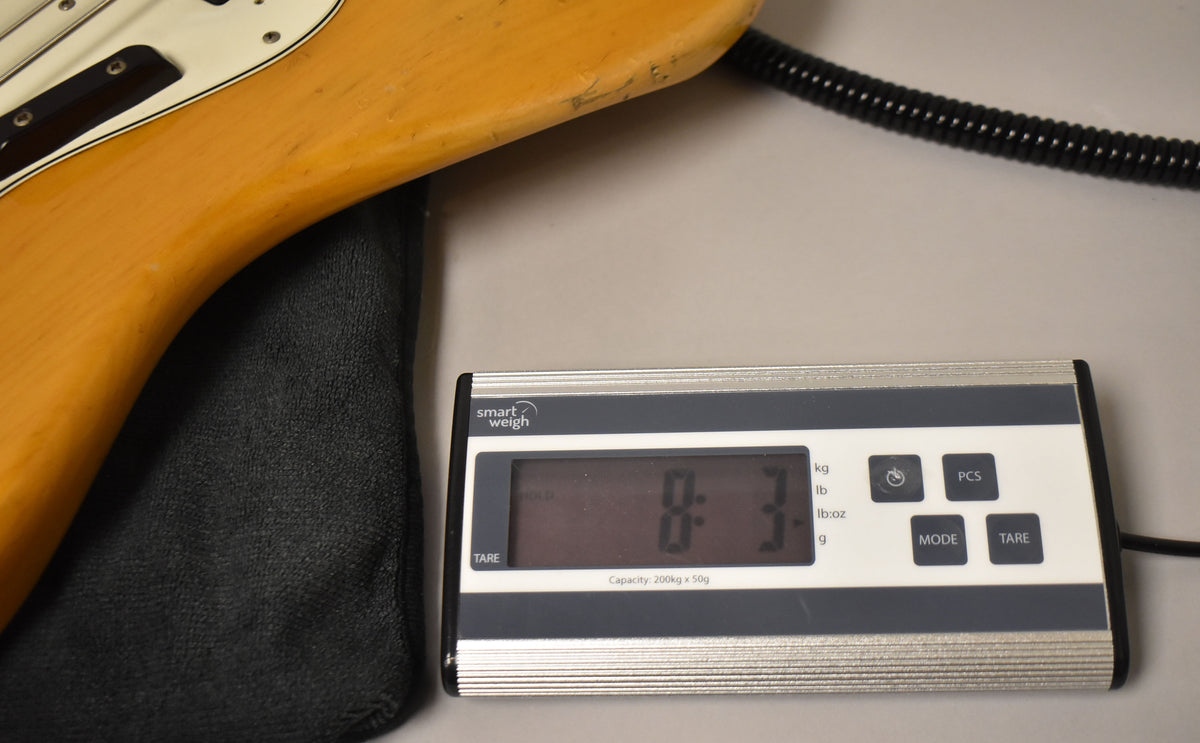 The natural finish bass body, with a white pickguard, rests on a black cloth and a digital scale displaying "8:3 lb".