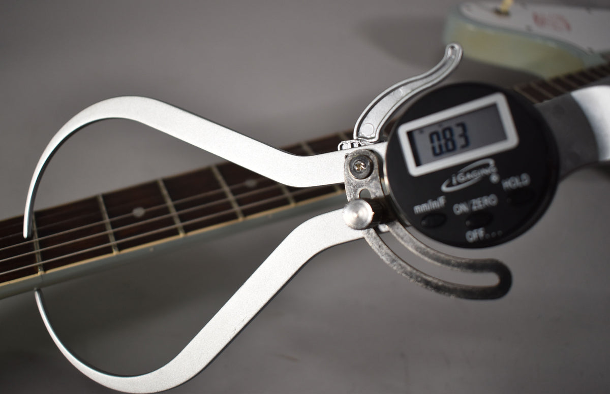 A digital caliper measures the guitar's fretboard, displaying "0.83" on its screen, with the light blue body visible in the background.