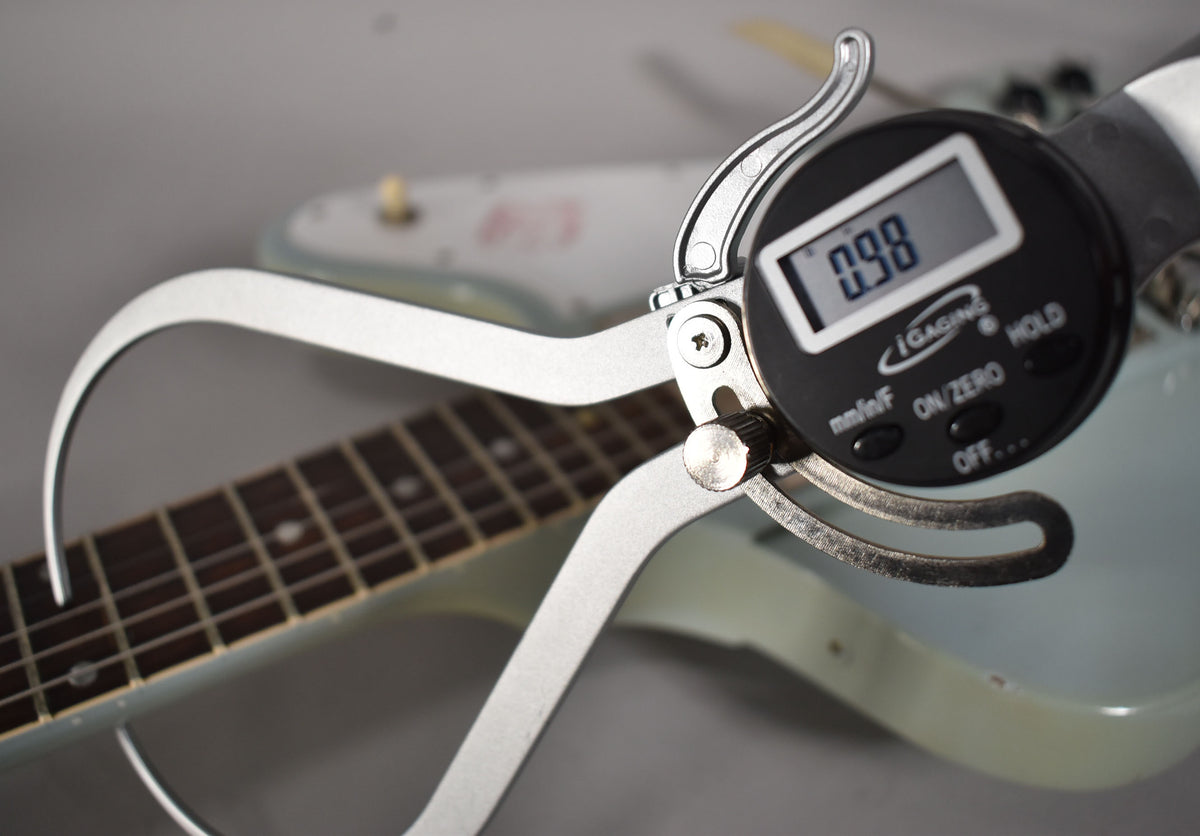 A digital caliper measures the guitar's fretboard, showing "0.98" on its screen, with the light blue body blurred behind.