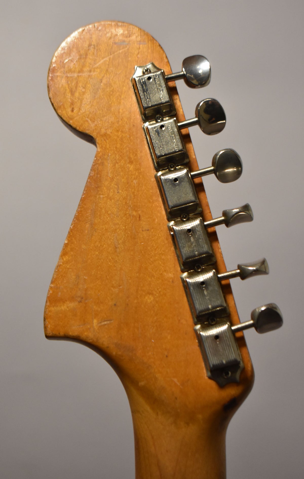 The headstock of a vintage electric guitar, angled left, shows six chrome tuners on a worn, natural wood finish.