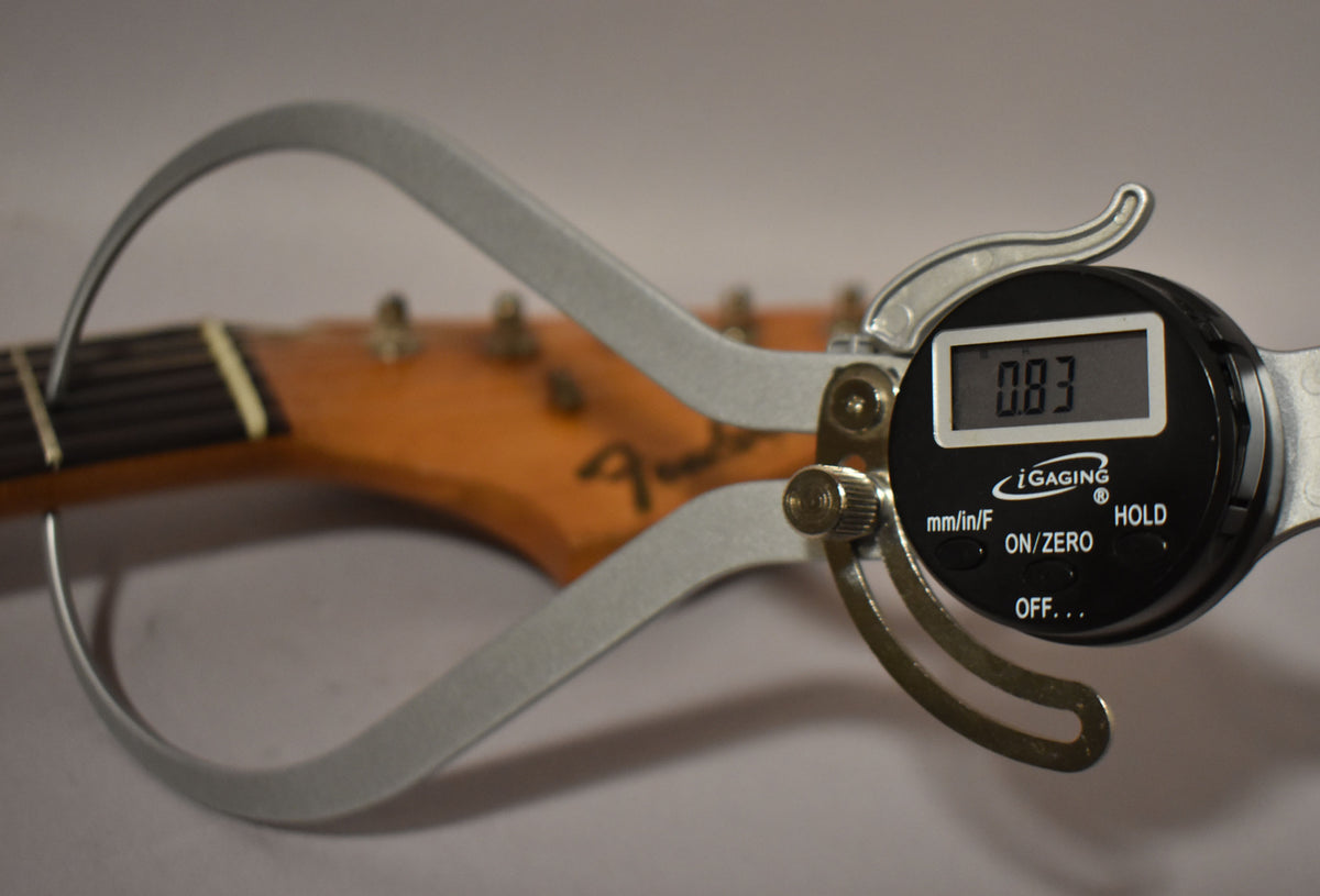 A digital caliper measures the neck thickness at the first fret, showing 0.83 inches, with the headstock and tuners visible.