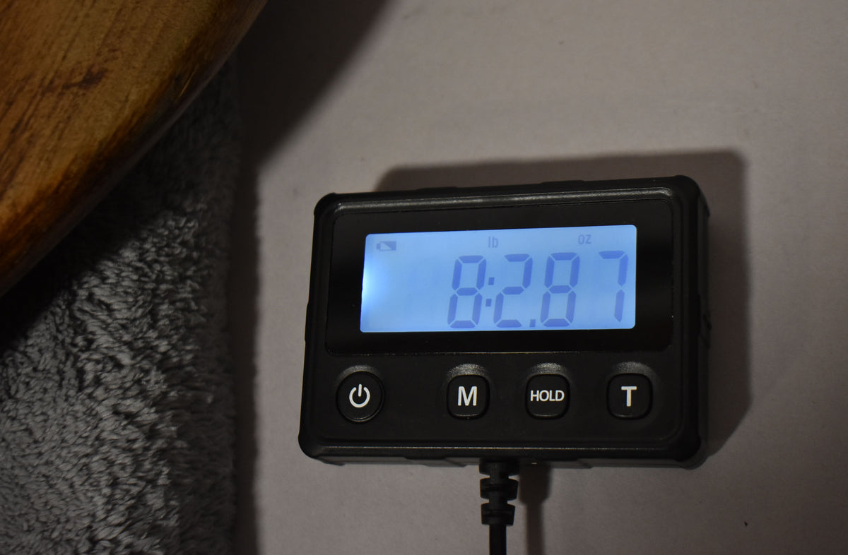 A close-up of a digital scale displaying "8:28.7" in blue, with a wooden object and gray fabric partially visible on the left.