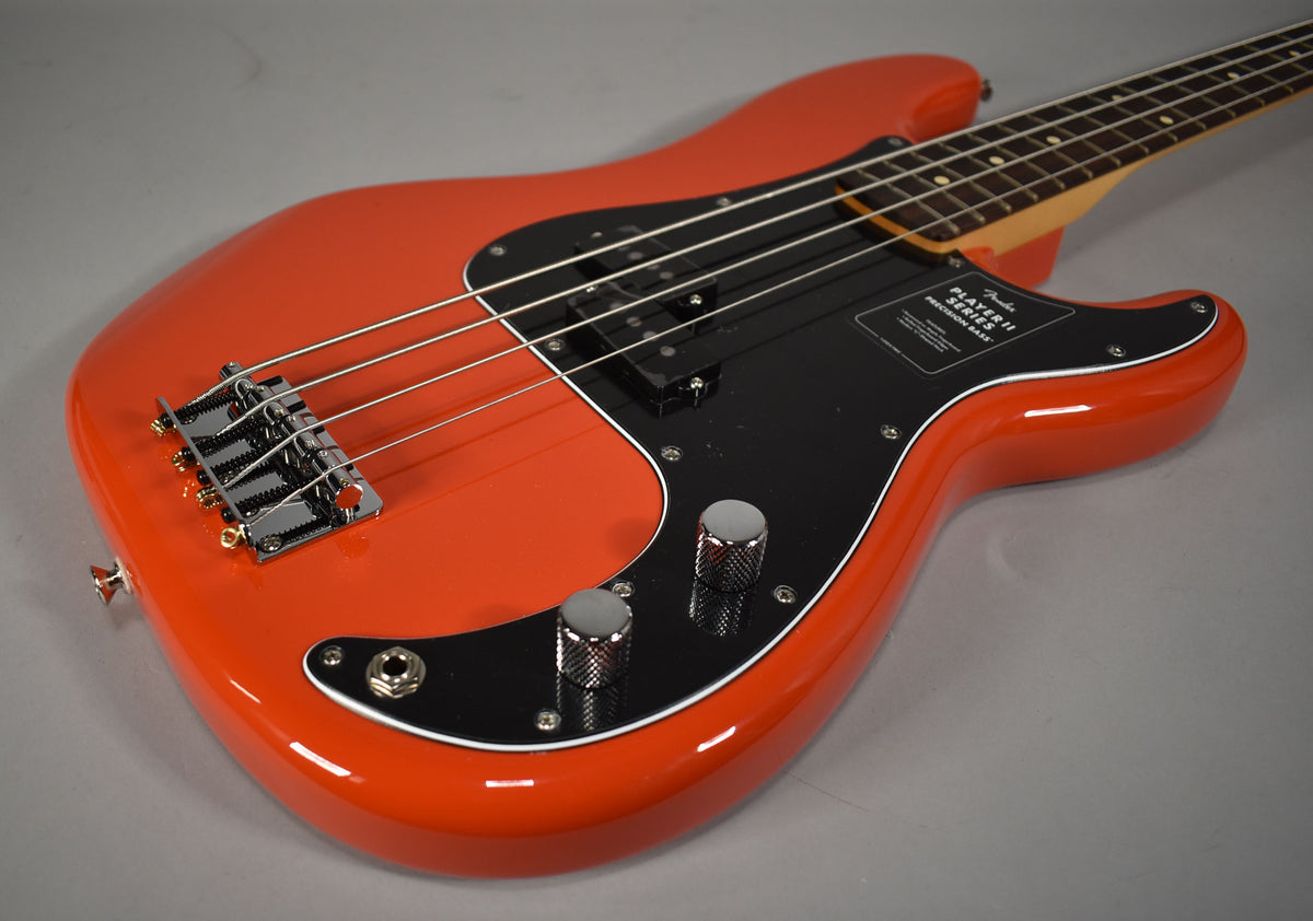 Coral red Precision Bass, angled from the lower bout, showing the glossy body, black pickguard, chrome bridge, and two knurled chrome control knobs.