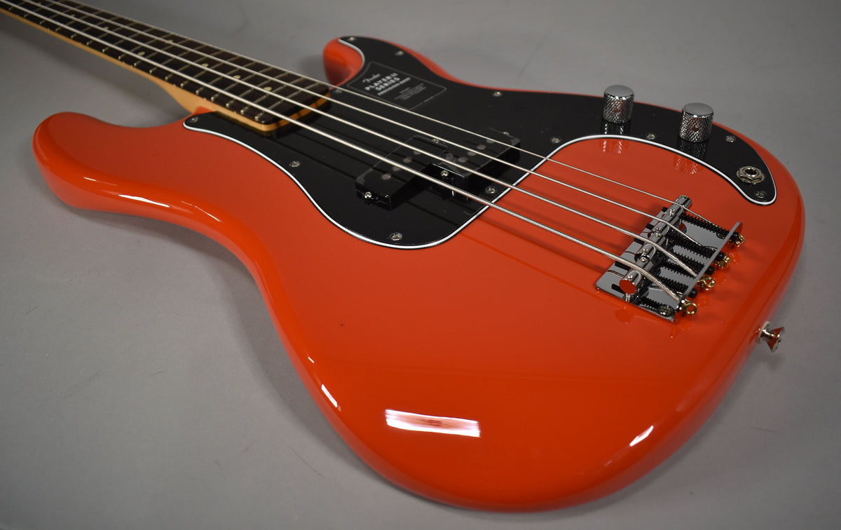 Coral red P-Bass, angled from the upper left, showing the glossy body, black pickguard, chrome bridge, and two knurled control knobs.