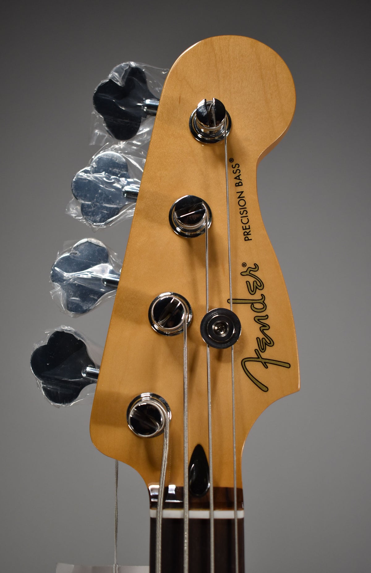 Close-up of the bass headstock, angled slightly right, showing four chrome tuners (three wrapped in plastic) and a black string tree.