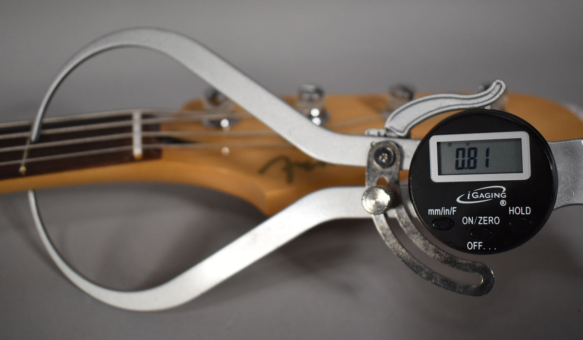 A close-up of the bass neck and headstock, with a digital caliper measuring the string height at 0.81.