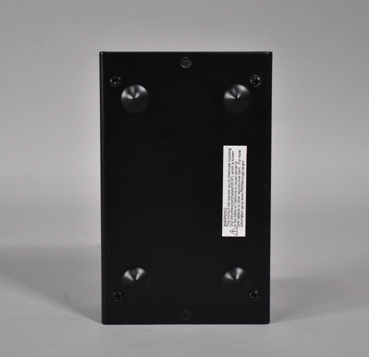 The black direct box is viewed from the bottom, showing four rubber feet and a white warning sticker on the smooth matte finish.