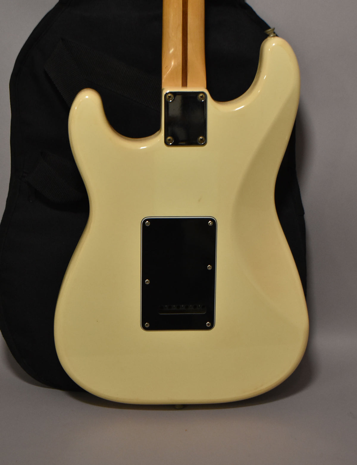 Rear view of an off-white Stratocaster body with a black tremolo cover and neck plate, showing a maple neck and a black gig bag.
