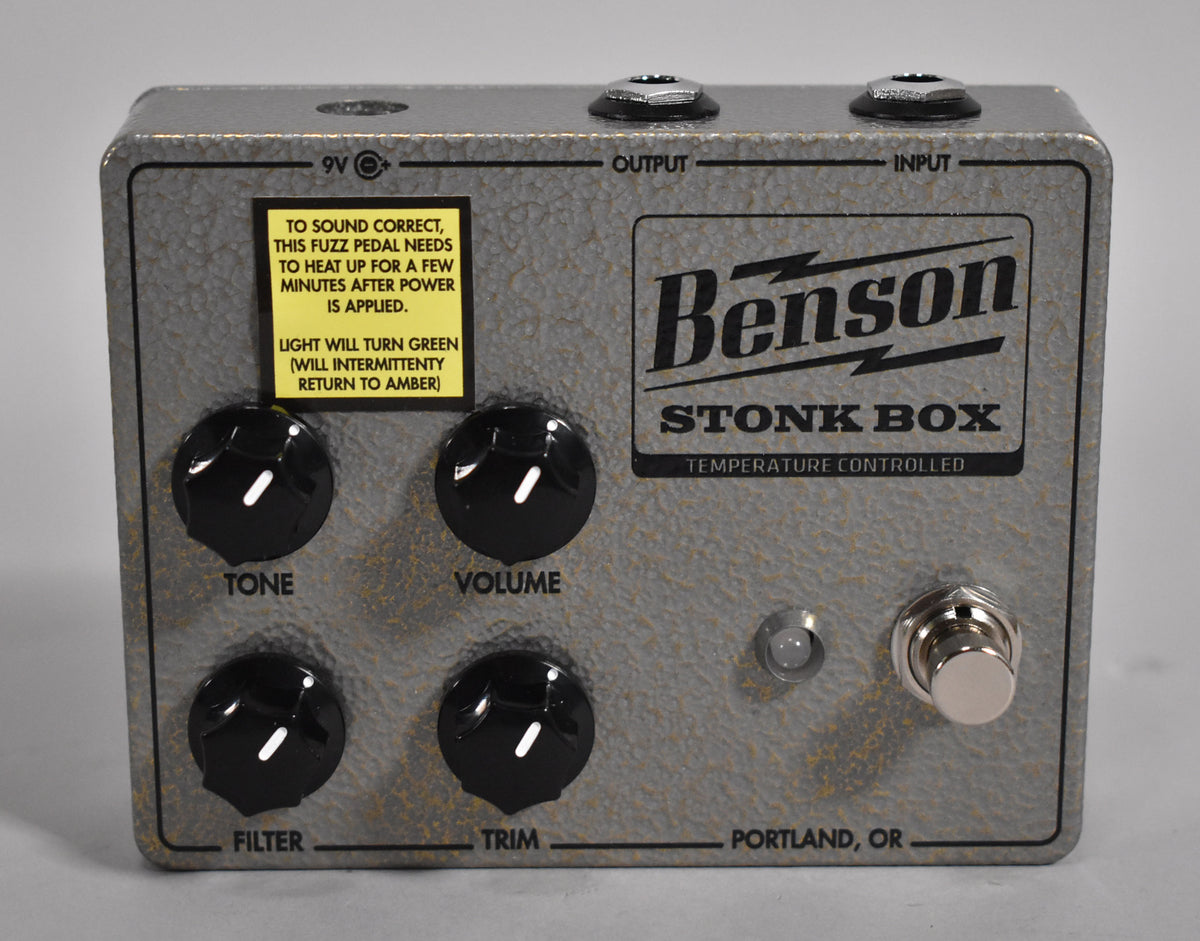 A gray textured fuzz pedal with four black knobs, a silver footswitch, and a small yellow warning sticker.