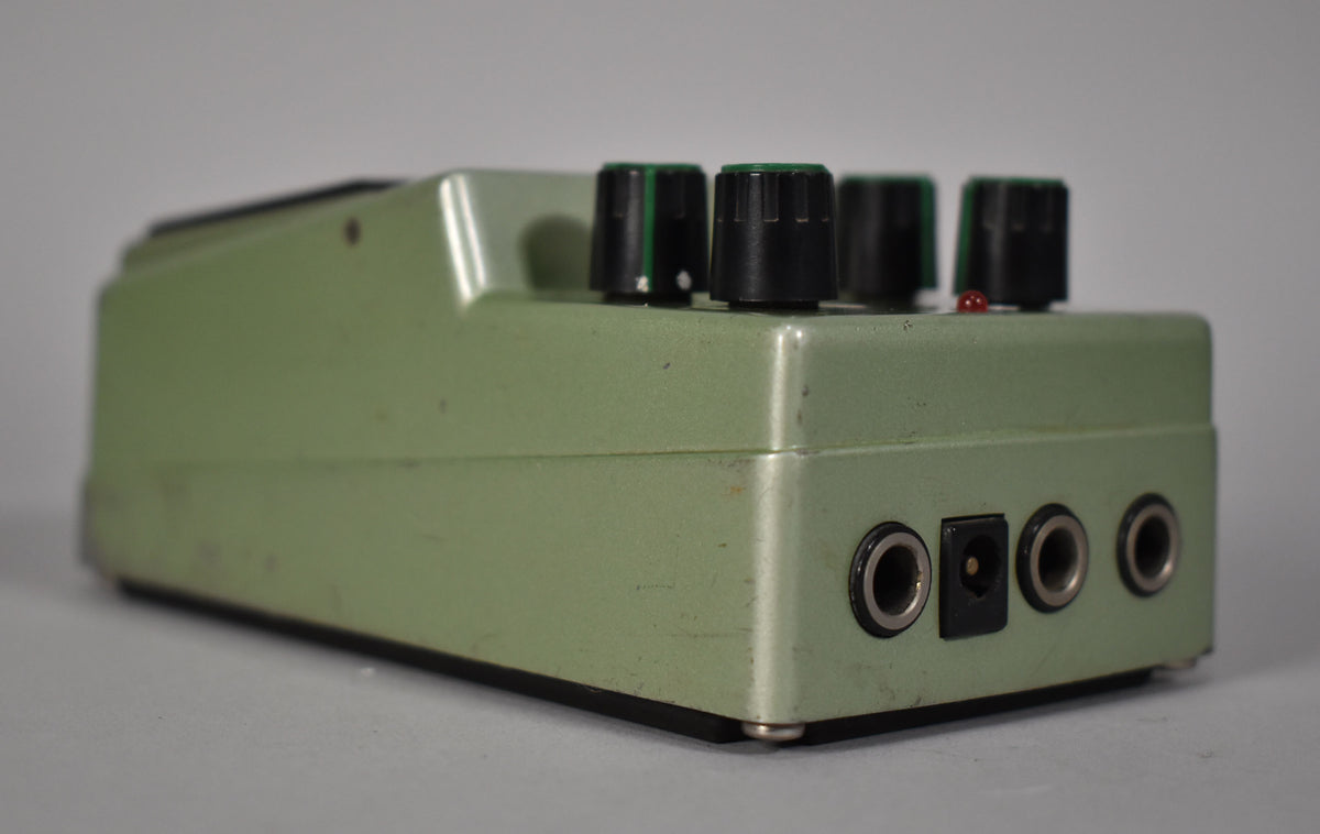 A pale green digital delay pedal, viewed from the right side, showing three black knobs with green bands, a red LED, and four jacks.