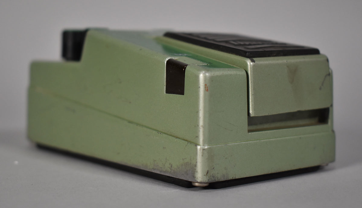 The green pedal is seen from the right side, showing a black rubber foot, a black input jack, and scuffs on the finish.