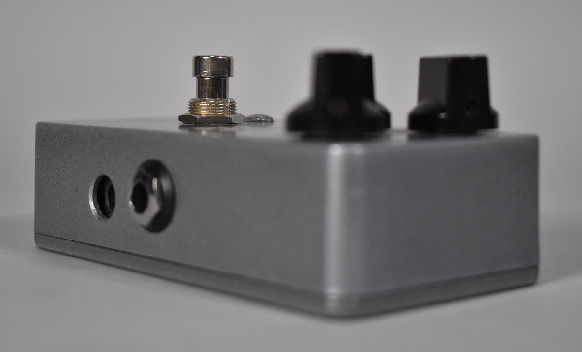 A silver-grey pedal, viewed from the left, showing two input jacks, a chrome footswitch, and two black control knobs.