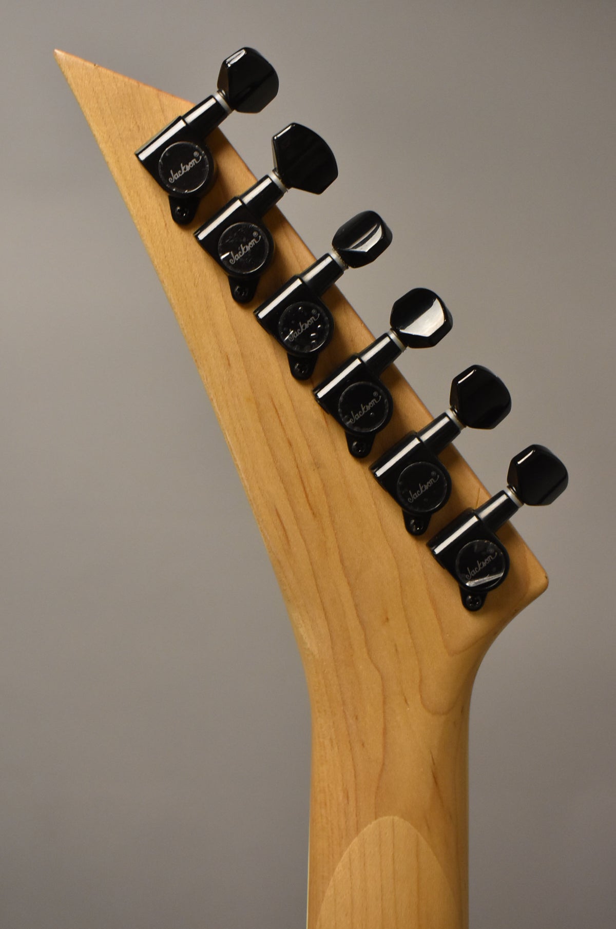 The back of a natural wood guitar headstock with six black tuning machines, angled from top left to bottom right.