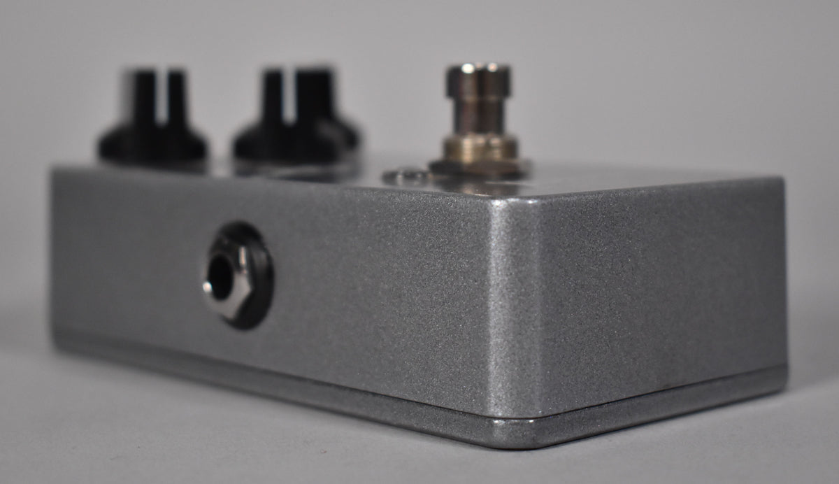 Side view of a metallic silver guitar pedal, showing two black control knobs, a silver footswitch, and an input jack.