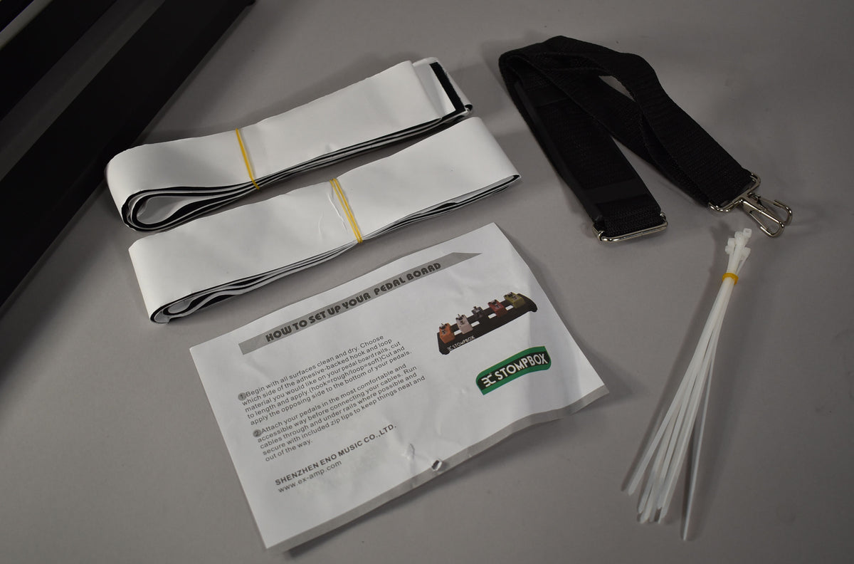 Overhead view of the pedalboard's accessories: two rolls of white hook-and-loop tape, a black shoulder strap, instructions, and a bundle of white zip ties.