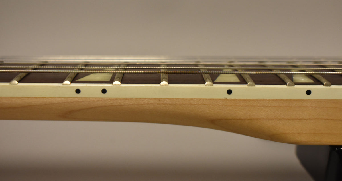 Close-up of the guitar's neck from the side, showing a light wood finish, dark fretboard with sharkfin inlays, and side dot markers.