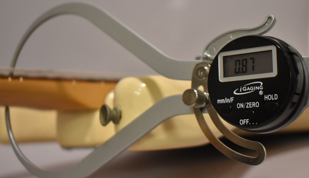 A digital caliper measures the Arctic White body and maple neck, displaying "0.87" on its screen.