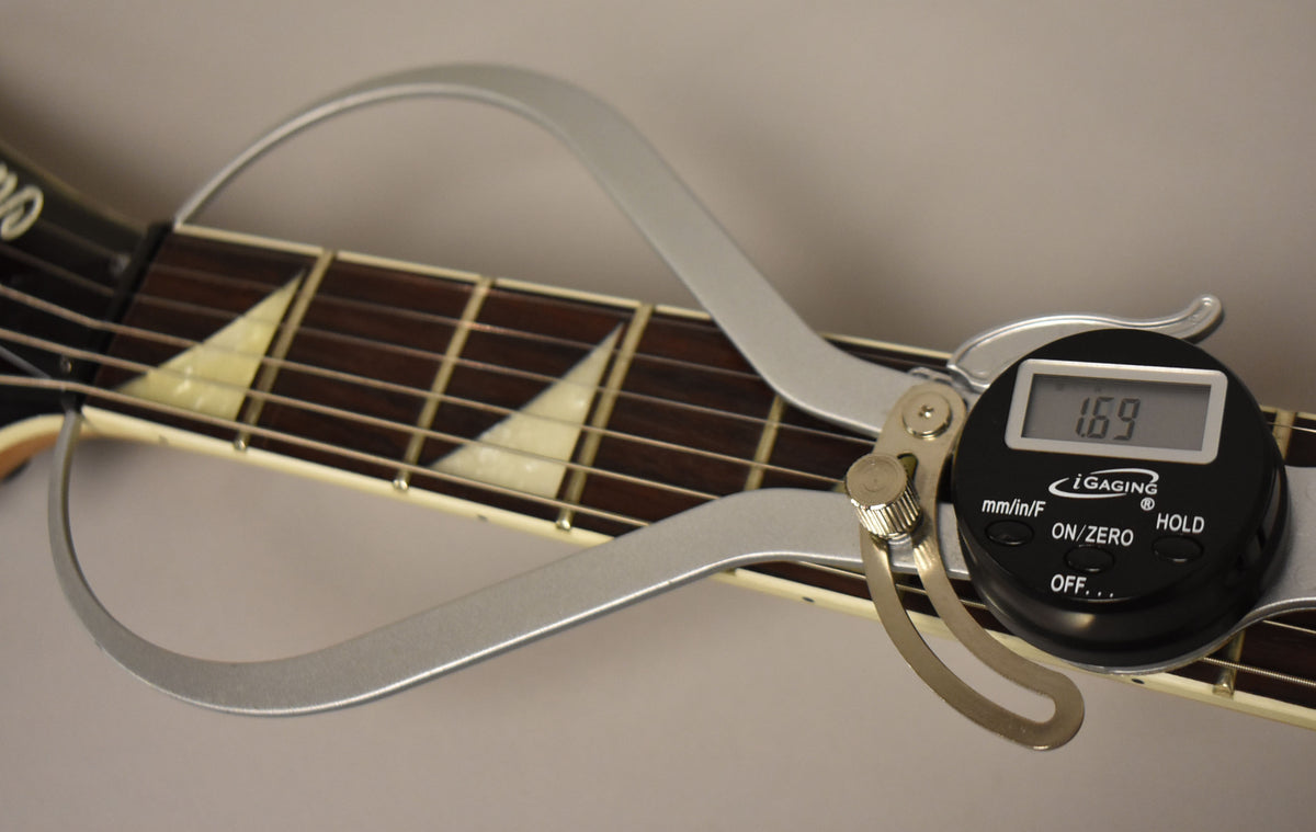 A digital caliper measures the string height at the 12th fret of the dark fretboard with white sharkfin inlays.