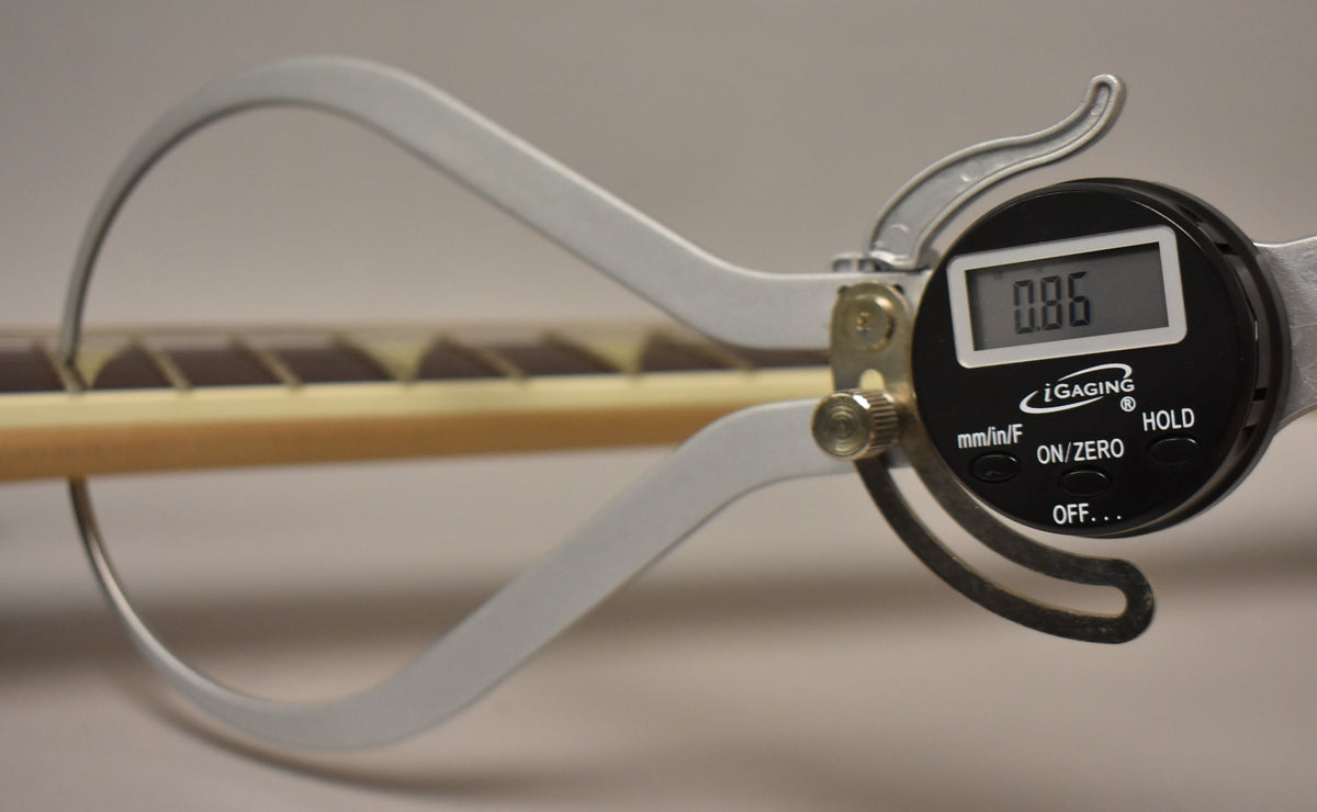A digital caliper measures the fret height of the guitar's light-colored neck, displaying "0.86" on its screen.