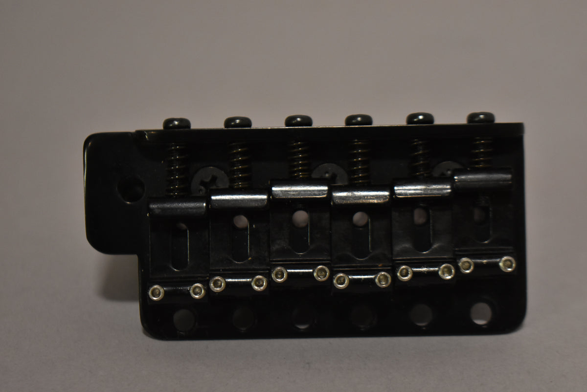 Black tremolo bridge, viewed from the side, showing six black saddles with silver-colored height adjustment screws and springs.