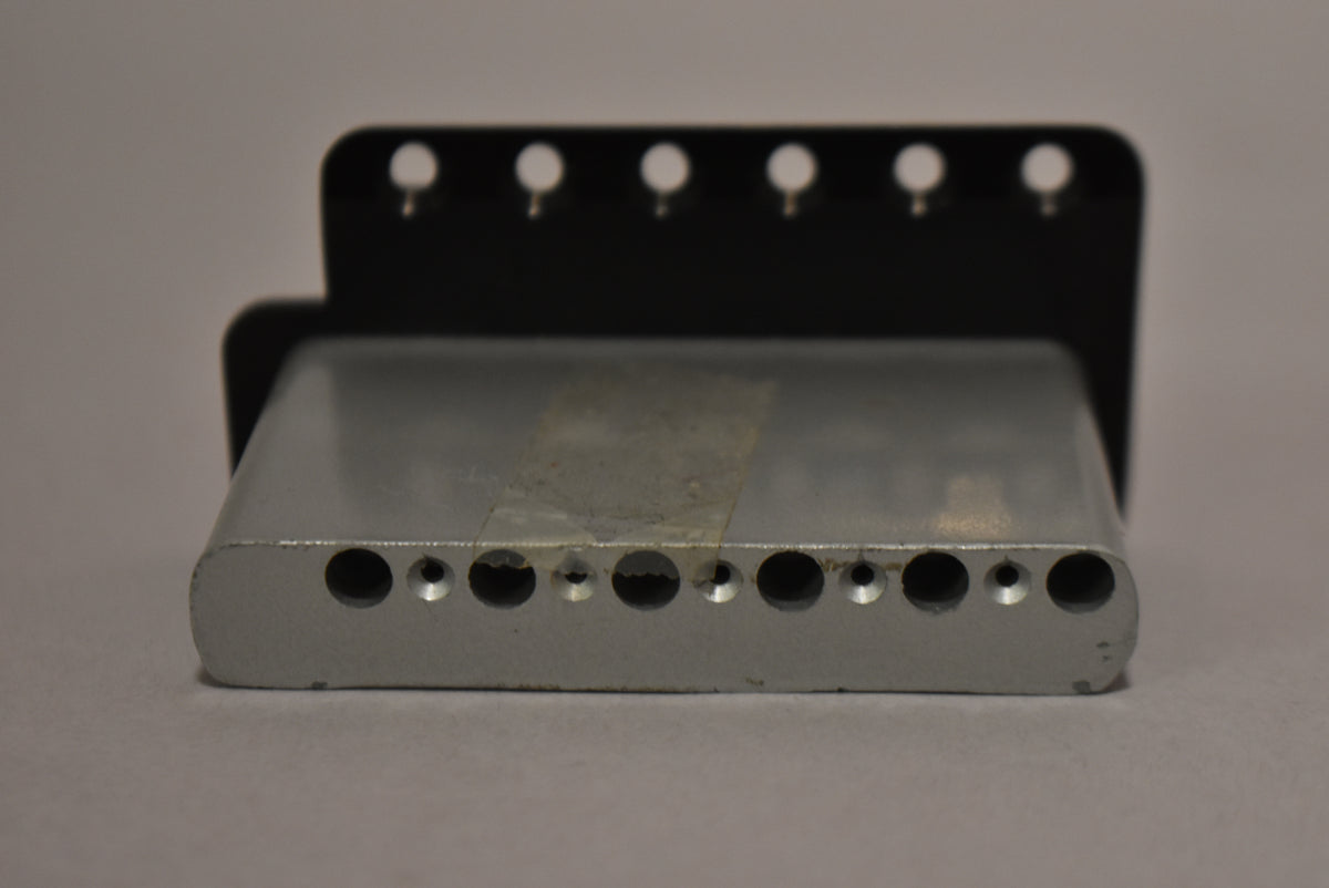 The black tremolo bridge is viewed from the rear, showing the silver block with six string holes and six mounting holes above.