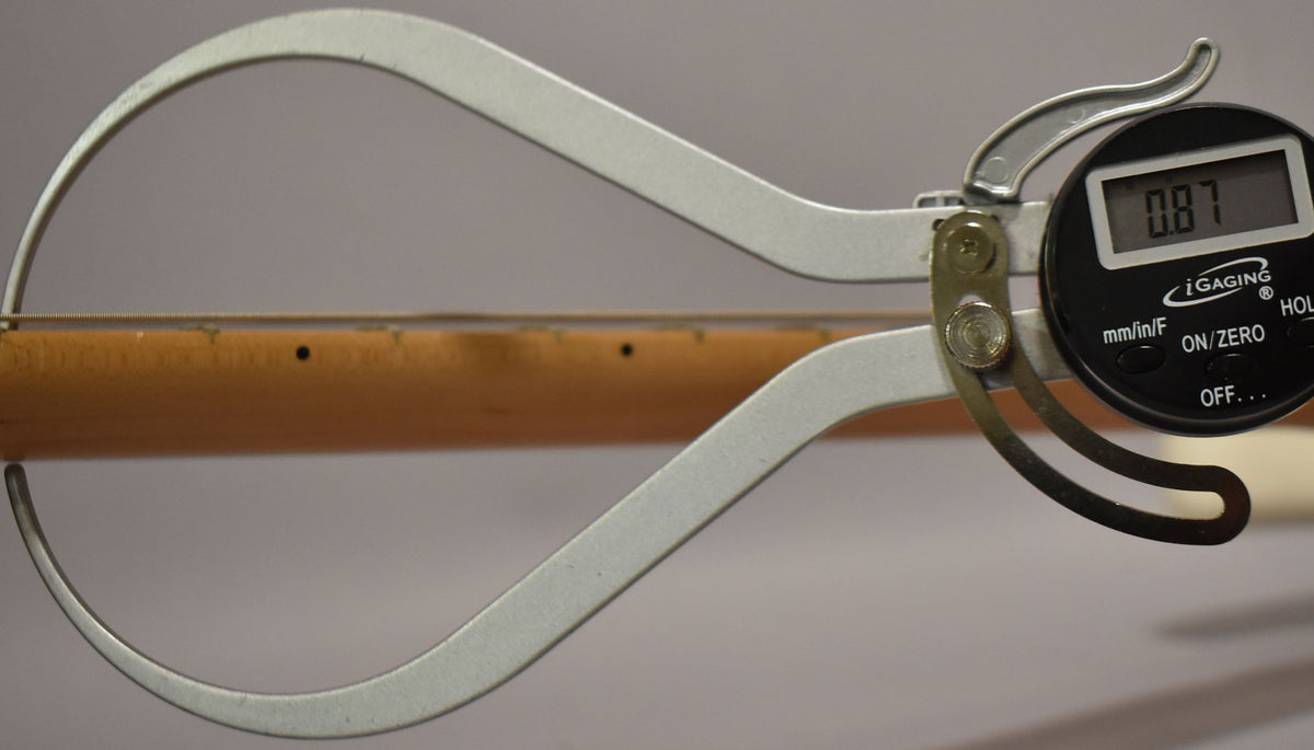 A digital caliper measures the maple neck of a white Stratocaster, showing a reading of 0.87.