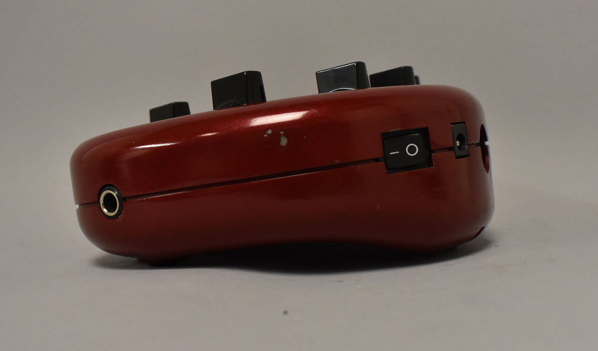 The red, kidney-bean-shaped pedal is shown from the side, revealing a power switch, input jack, and some paint chips.