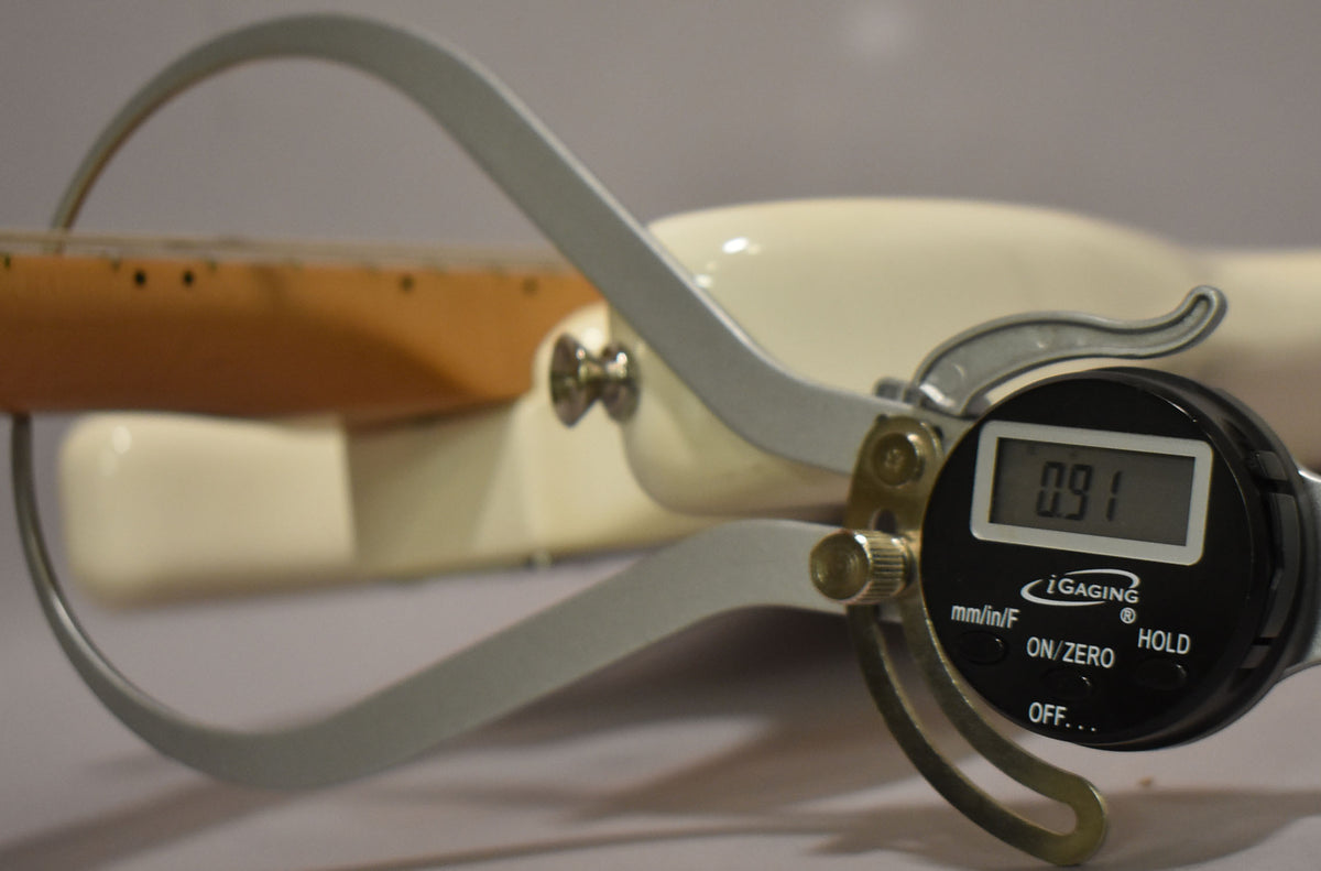 A digital caliper measures the neck thickness of the polar white Stratocaster, displaying "0.91" on its screen.