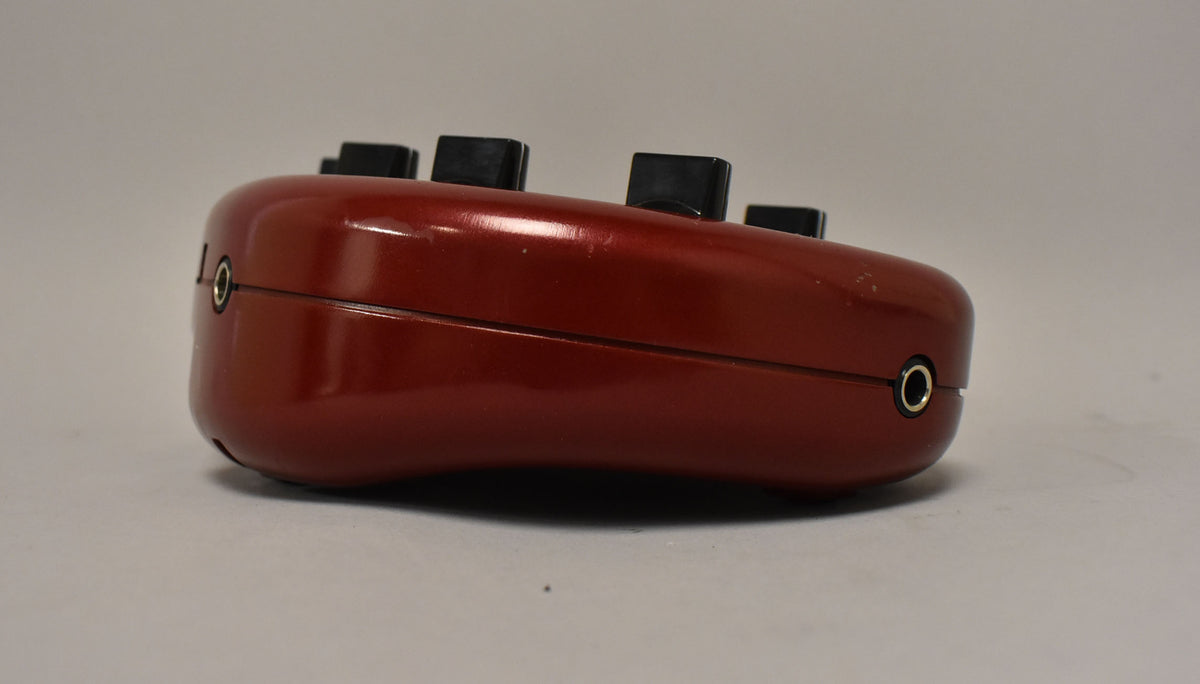 The red, kidney-bean-shaped pedal is viewed from the side, showing two 1/4-inch jacks and several black control knobs on top.
