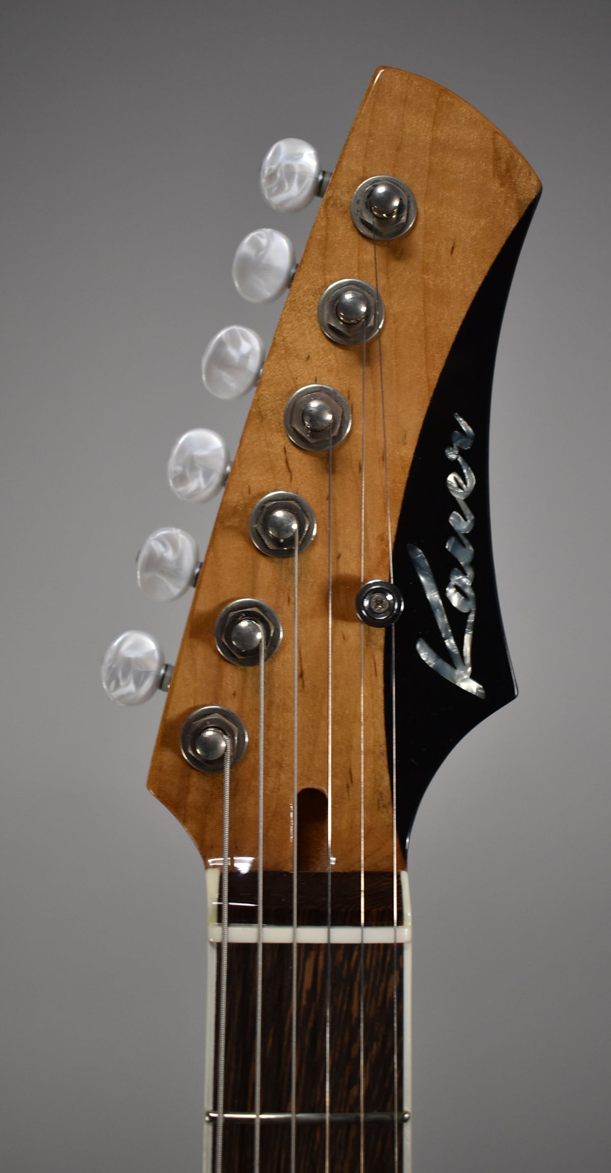 Close-up of the guitar's headstock, showing a natural wood finish, six mother-of-pearl tuners, and a black headstock face with an inlaid logo.